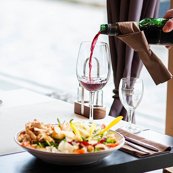 Eine Schüssel mit einen zubereitetem Essen und Gemüse, eine Hand füllt Wein in ein Weinglas daneben.
