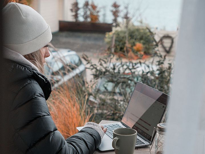 PErson in Winterkleidung auf Terrasse mit Laptop, im Hintergrund blauer See