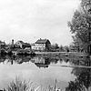 Schwarz-Weiß-Foto eines Sees. Hinter dem See stehen einige Häuser. Das rechte Ufer ist gesäumt mit Bäumen. 