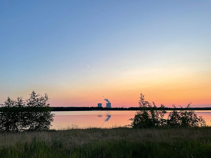 Grünes Ufer vor einem See im Sonnenuntergang, am Horizont erkennt man zwei kleine Kühltürme.