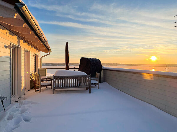 Dachterrasse mit Schnee im Sonnenuntergang über dem Hainer See. Am Ende der Terrasse stehen eine Sitzbank, ein Tisch mit Sesseln, ein Sonnenschirm und ein Strandkorb.