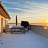 Dachterrasse mit Schnee im Sonnenuntergang über dem Hainer See. Am Ende der Terrasse stehen eine Sitzbank, ein Tisch mit Sesseln, ein Sonnenschirm und ein Strandkorb.