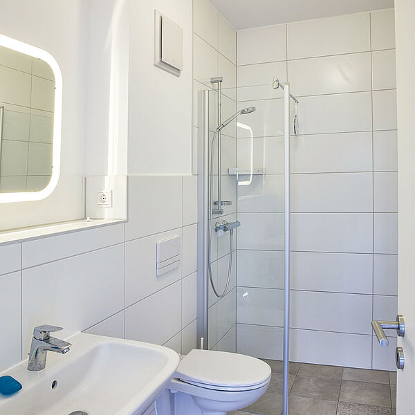  Bad mit ebenerdiger Dusche. Daneben eine Toilette und ein Waschbecken mit beleuchtetem Spiegel.