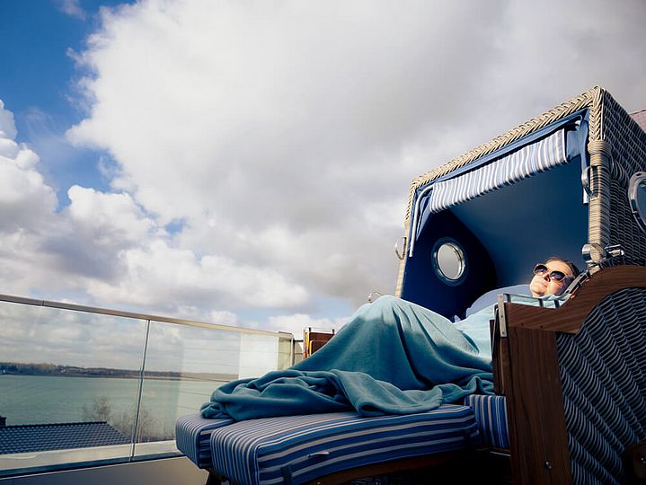 Eine Person mit Sonnenbrille liegt eingekuschelt in einer Decke auf einem Strandkorb für zwei Personen in der Sonne. Durch das Glasgeländer der Dachterrasse sieht man den See.