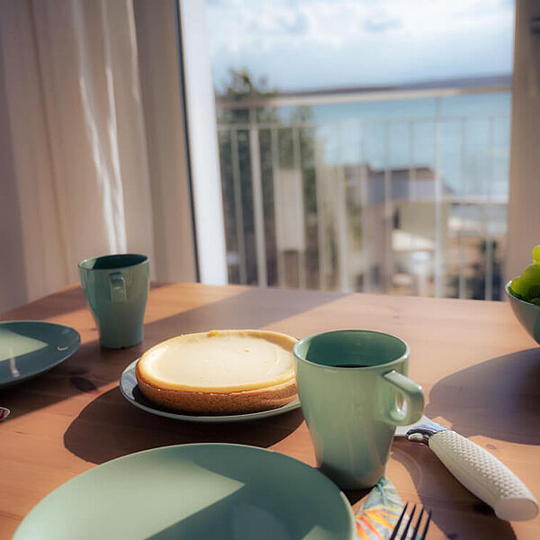 Gedec, Käsekuchen und Trauben auf dem Esstisch, im Hintergrund der türkise Hainer See.