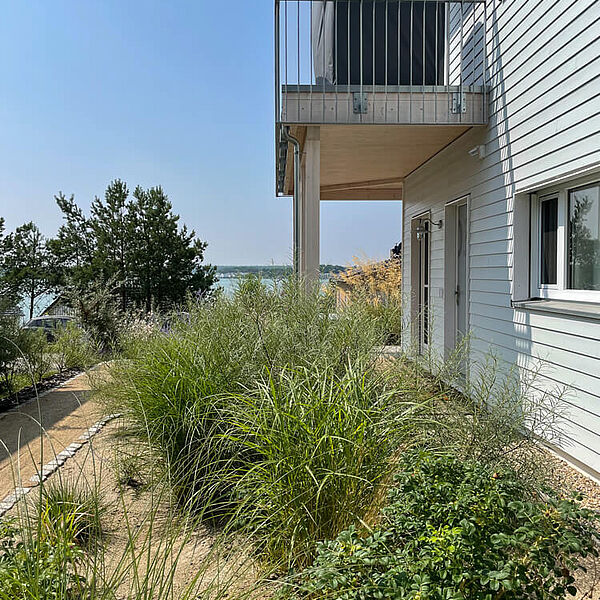 Sommerlicher Garten mit Blick auf den Hainer See. Rechts eine Hauswand mit Terrasse und darüberliegendem Balkon.