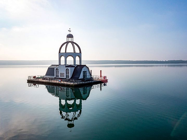 Mitten im Wasser ragt eine schwimmende Insel in Form eines Kirchturms aus dem Wasser.