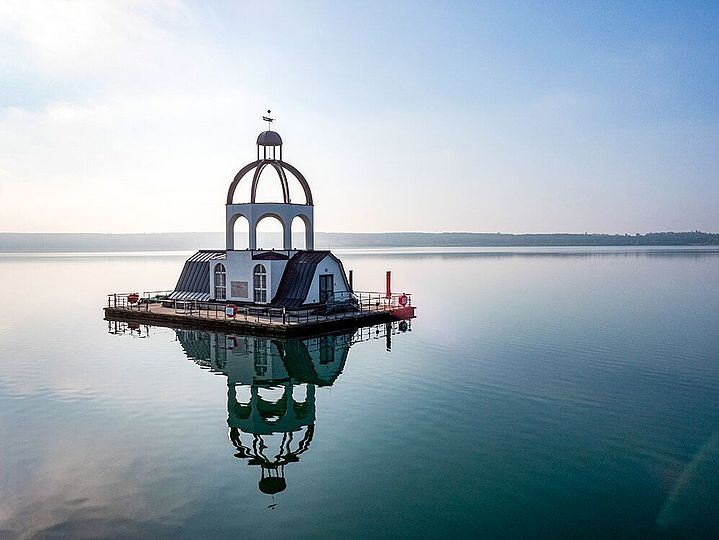 Mitten im Wasser ragt eine schwimmende Insel in Form eines Kirchturms aus dem Wasser.
