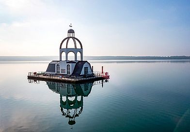 Mitten im Wasser ragt eine schwimmende Insel in Form eines Kirchturms aus dem Wasser.