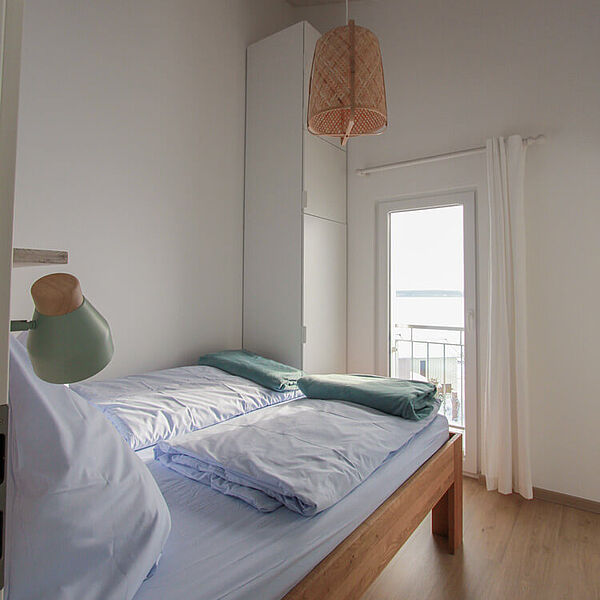 Doppelbett aus Holz. Gegenüber ein bodentiefes Fenster mit weißen Vorhängen an den Seiten.