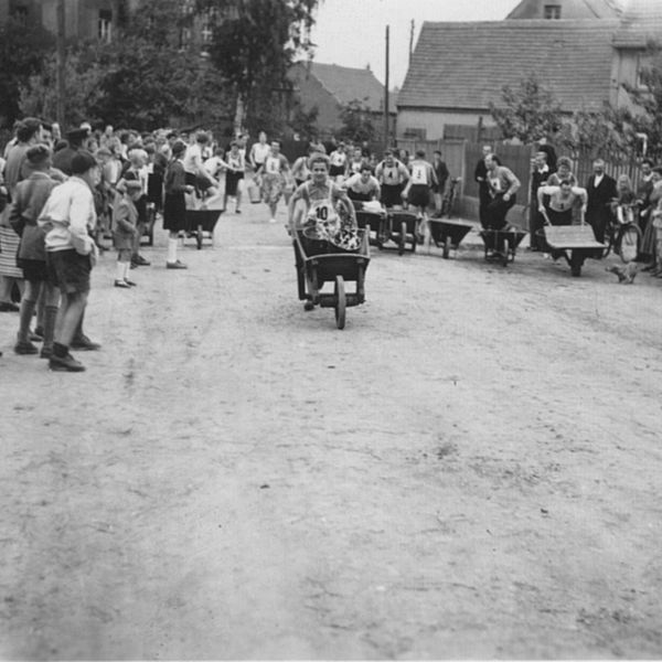Schwarzweiß-Foto einer Dorfstraße mit Menschen an den Seiten, die bei einem Schubkarrenrennen zuschauen.