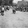 Schwarzweiß-Foto einer Dorfstraße mit Menschen an den Seiten, die bei einem Schubkarrenrennen zuschauen.